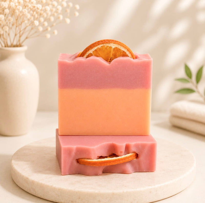 Four bars of Mimosa handcrafted artisan bar soap stacked on a round wooden tray. The soap bars are pink with a peach-colored side and a dried orange slice embedded in the top, set against a softly blurred background with a vase of dried flowers.