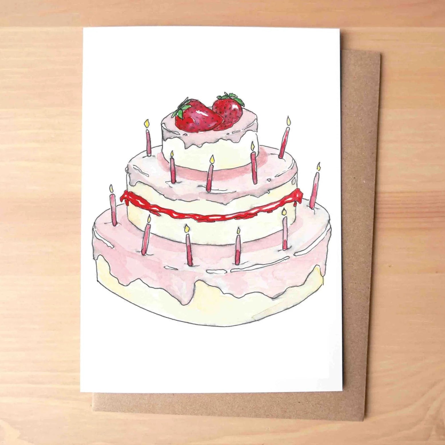 A white greeting card featuring a watercolor illustration of a three-tiered birthday cake with candles and strawberries, placed on a brown kraft envelope over a light wood tabletop.