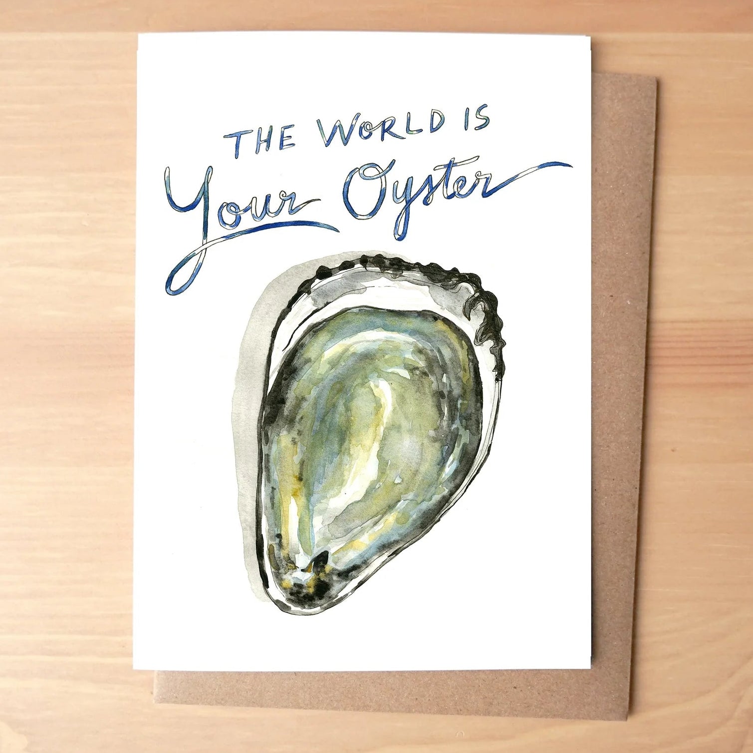 A white greeting card with the phrase 'THE WORLD IS Your Oyster' in blue script and a watercolor illustration of an oyster shell, placed on top of a brown kraft envelope over a light wood surface.