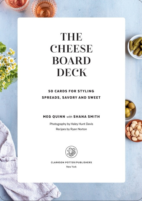 The Cheese Board Deck card game box by Meg Quinn, featuring a rustic wooden board design filled with cheeses, grapes, cured meats, crackers, and citrus slices. The front displays the title 'THE CHEESE BOARD DECK' 