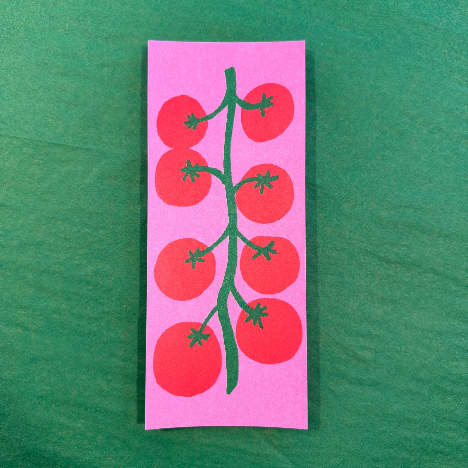 A rectangular pink notepad featuring a stylized green vine with red tomatoes, placed on a green background.