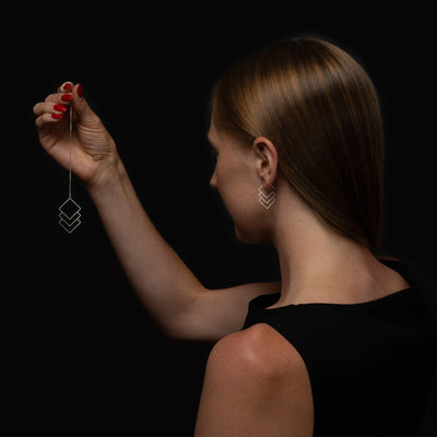 A woman in a black top holds up a delicate silver geometric dangle earring against a solid black background.