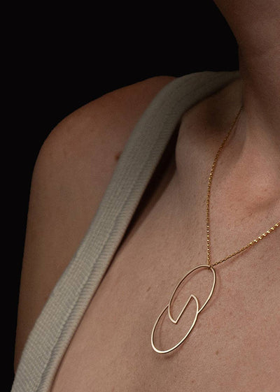 A close-up of a model wearing a delicate gold necklace with a large, open interlocking oval pendant. The model is wearing a light beige knit top against a solid black background.
