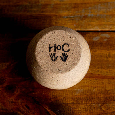 Round ceramic match holder with 'HoC' and hand symbols on a wooden surface