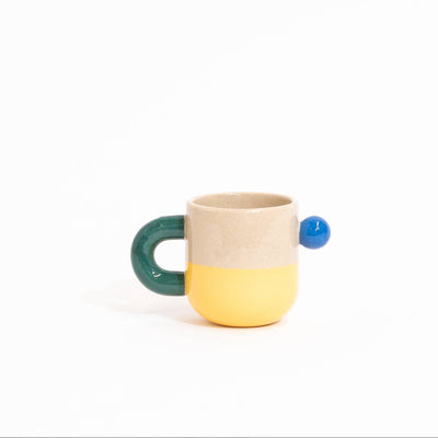 A small ceramic cup with a split beige and yellow body, a dark green handle, and a small blue accent on the side, set against a plain white background.