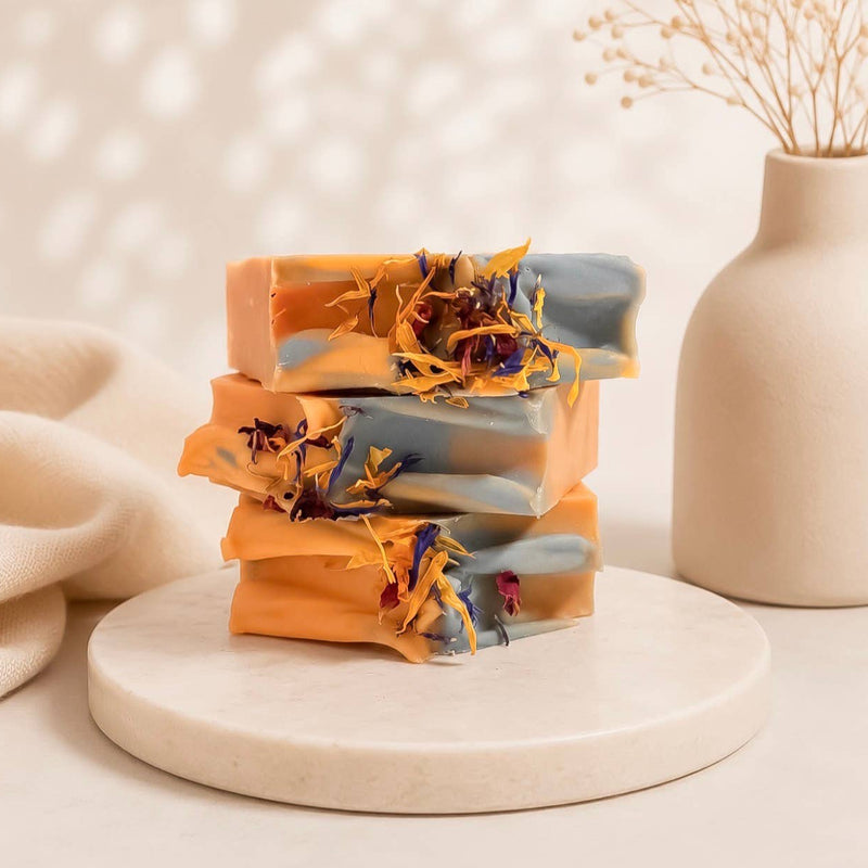 A handmade artisan bar soap labeled 'PASSIONFRUIT & PATCHOULI' by Spirit Glow Home & Body Essentials, stacked on a white marble tray with dried botanicals, set against a soft beige background with a ceramic vase.