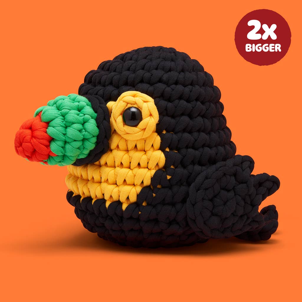 A crocheted plush bird toy with a black body, yellow chest, green & orange  beak, and black eye, shown on an orange background with a red '2x BIGGER' badge in the top right.