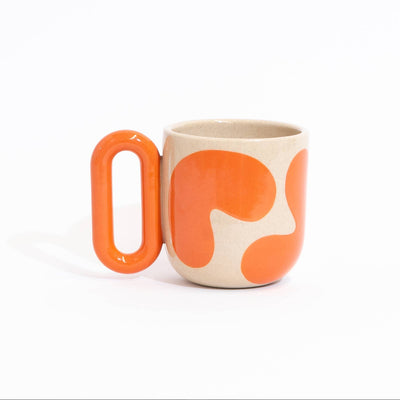A ceramic mug with a bold orange abstract pattern on a warm cream background, featuring a unique rounded rectangular handle, set against a plain white studio background.