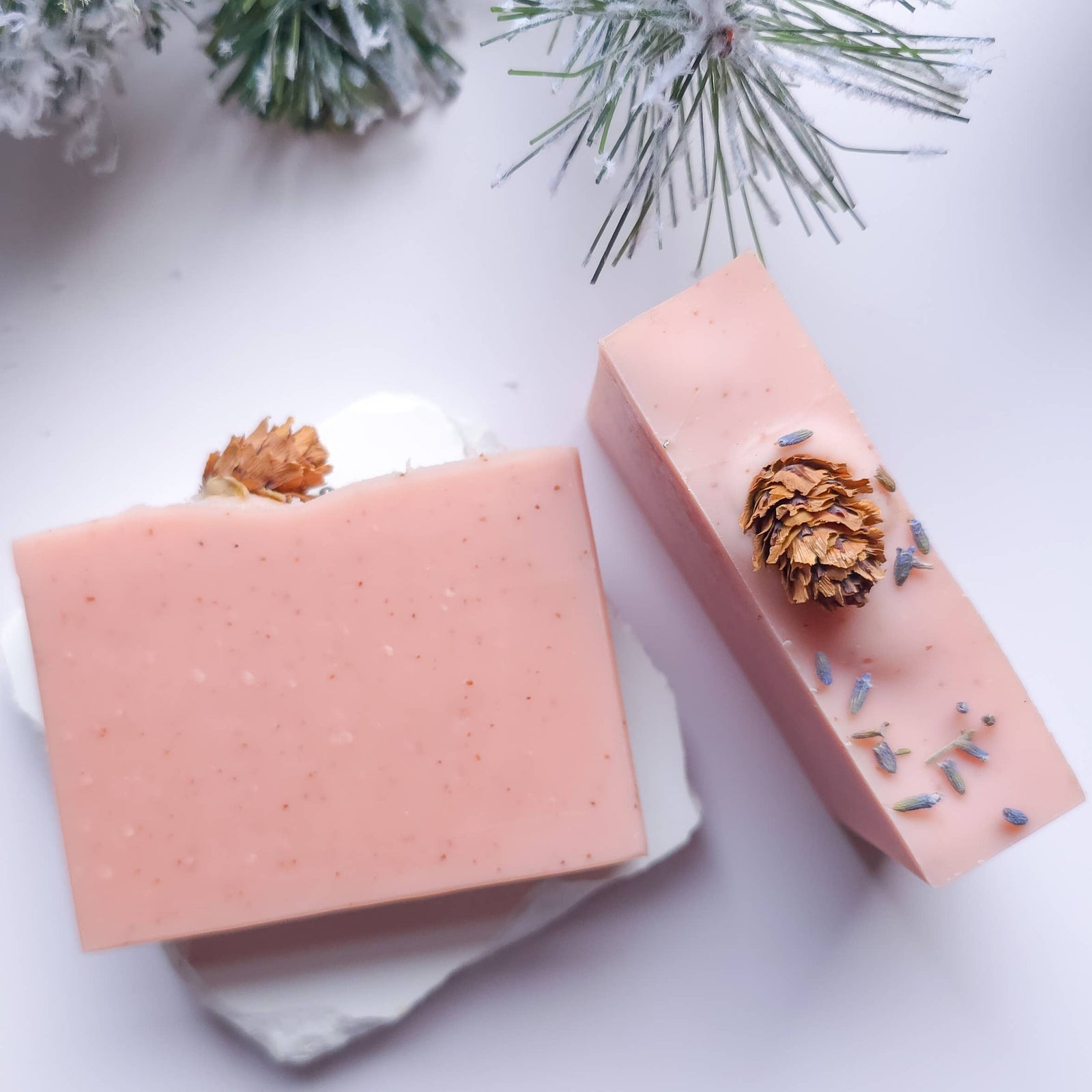 Two pink soap bars with decorative elements on a white surface with snow and pine branches.