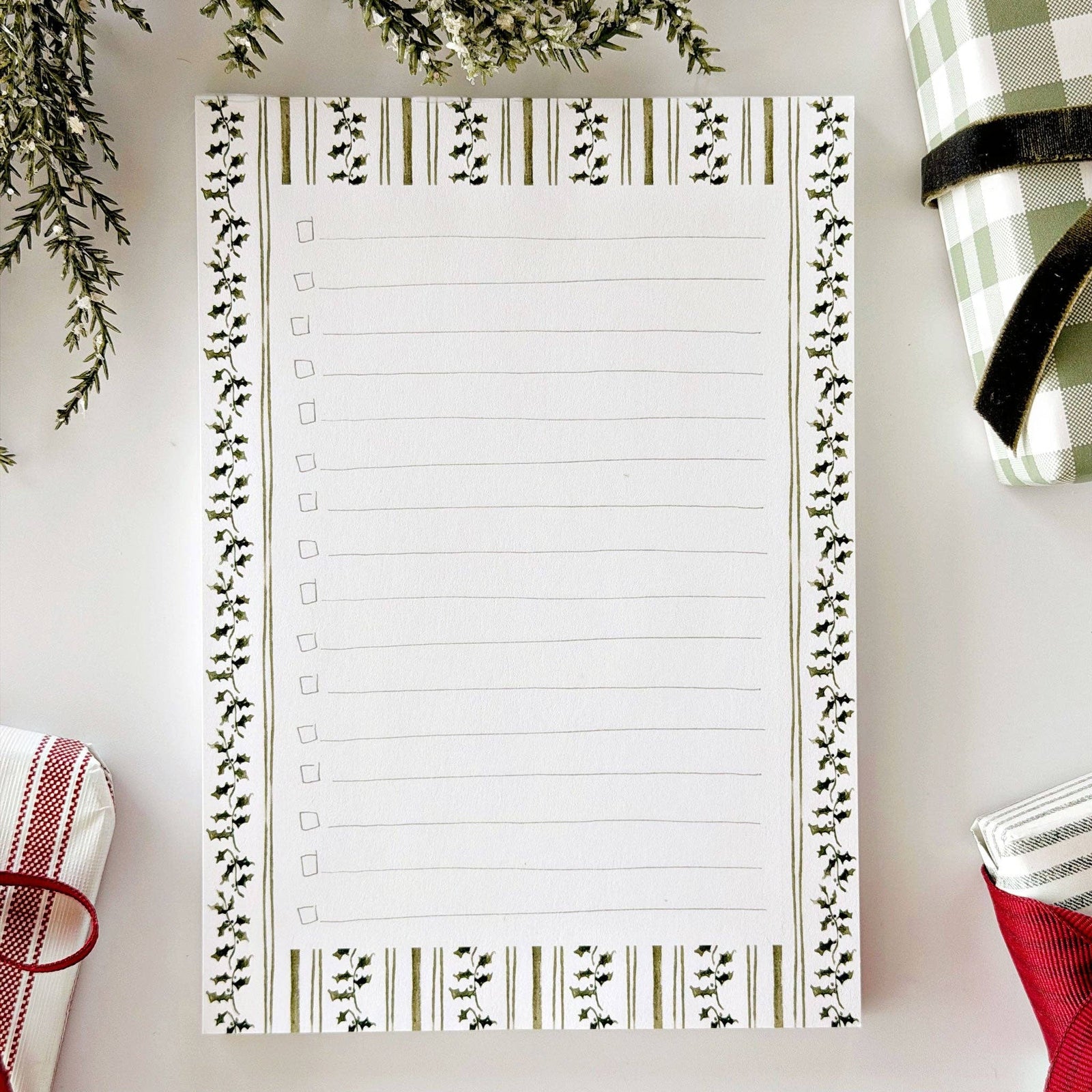 A lined notepad with a decorative border of green leaves and gold stripes, laid flat on a white surface with holiday-themed props around it.