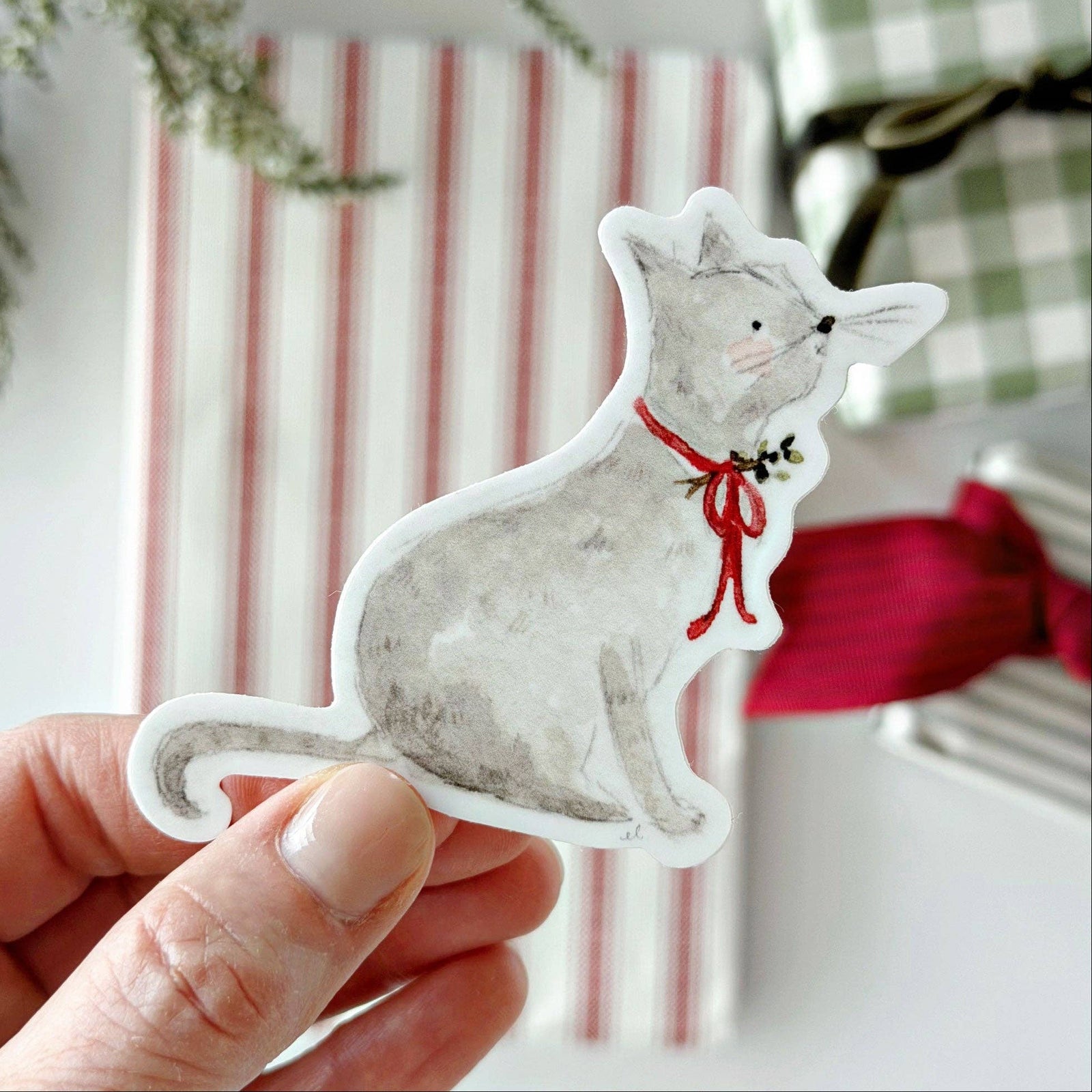 A hand holding a sticker of a cat with a red bow on a light blurry background.