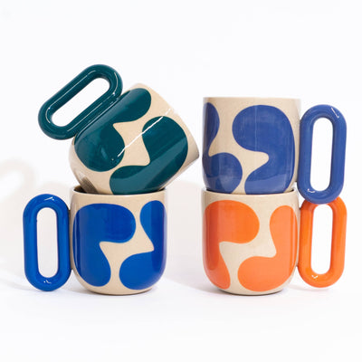 Three ceramic mugs with bold abstract color-blocked patterns in blue, orange, and teal on a cream base, arranged on a clean white background.