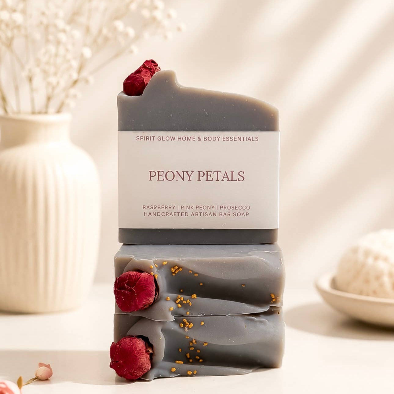 A stacked set of handmade artisan soap bars in a warm neutral setting. The soaps are a muted grayish-brown with a white label reading 'PEONY PETALS' and decorated with fresh red raspberries and gold flakes.