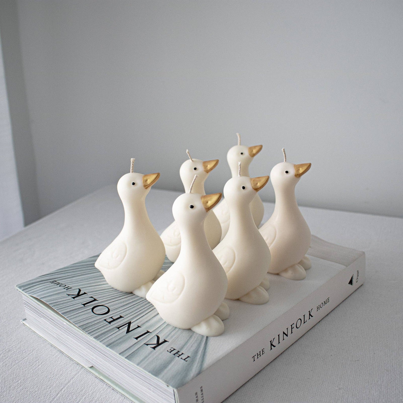 Six white goose shaped candles with gold beaks are arranged on top of a  book against a light gray background.