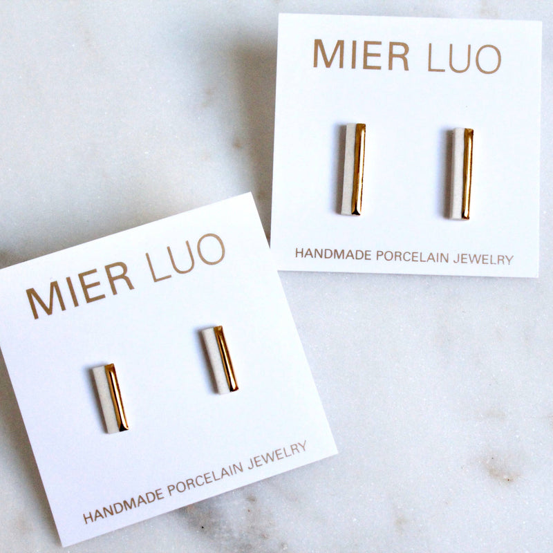 A pair of minimalist gold and  bar stud earrings resting on a light marble surface.