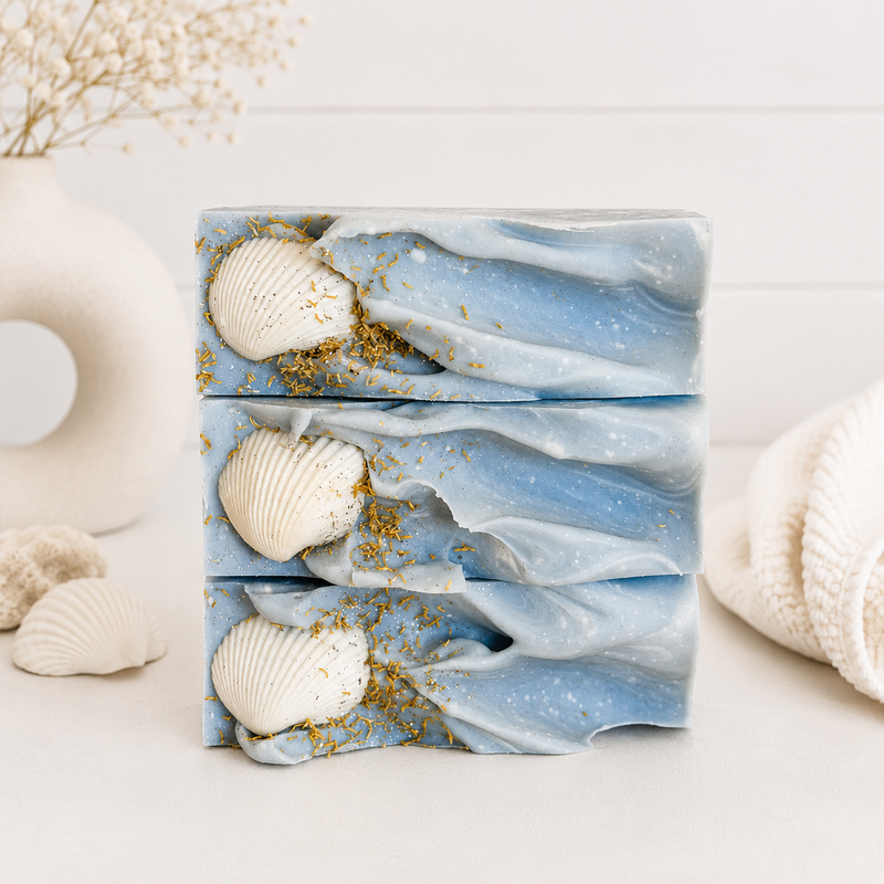 A handmade artisan bar soap labeled 'COASTAL WAVES' by Spirit Glow Home & Body Essentials, stacked with seashells and gold flakes on a light surface with a soft beach-themed background.