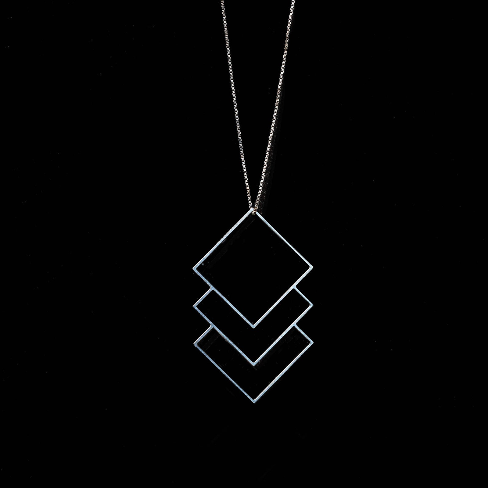 A silver geometric pendant necklace featuring three stacked diamond outlines on a fine chain, set against a solid black background.