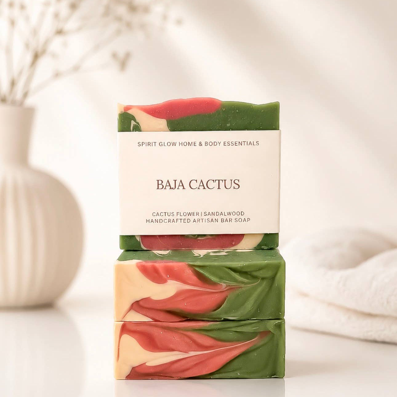 Two stacked handmade artisan bar soaps labeled 'BAJA CACTUS' by Spirit Glow Honey & Body Essentials, featuring green, coral pink, and cream swirls, set against a soft, warm, blurred background with a white vase and towel.