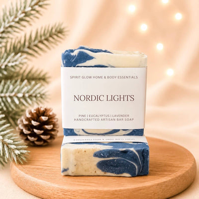 A handmade artisan bar soap labeled 'Nordic Lights' by Spirit Glow Home & Body Essentials, displayed on a round wooden board with a second soap piece showing its marbled blue and cream texture. The scene is decorated with a pine cone, evergreen branches, and warm bokeh string lights in the background.