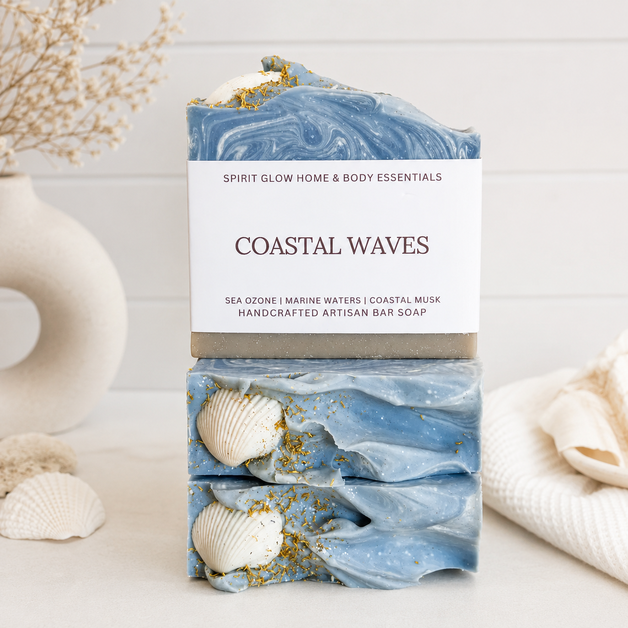A handmade artisan bar soap labeled 'COASTAL WAVES' by Spirit Glow Home & Body Essentials, stacked with seashells and gold flakes on a light surface with a soft beach-themed background.