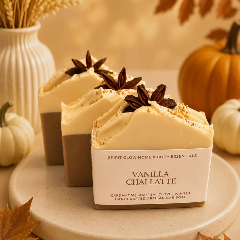 A bar of Vanilla Chai Latte artisan soap displayed on a ceramic plate, with its branded paper sleeve behind it. The soap is cream-colored, set against a warm autumnal background with pumpkins, dried grass, and leaves.