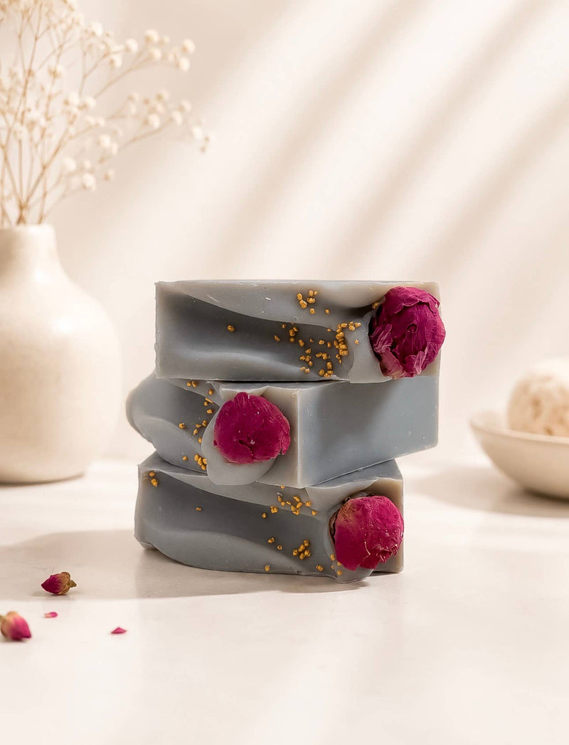 A stacked set of handmade artisan soap bars in a warm neutral setting. The soaps are a muted grayish-brown with a white label reading 'PEONY PETALS' and decorated with fresh red raspberries and gold flakes.