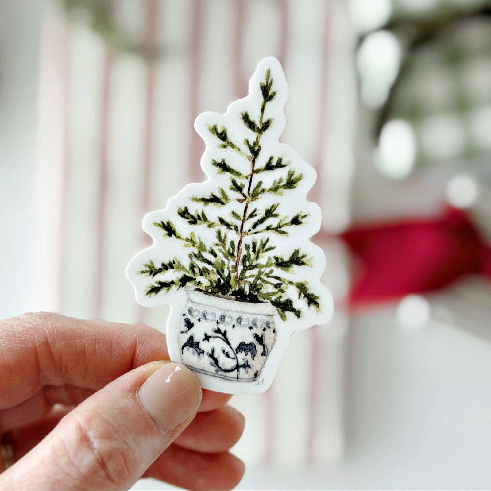 A hand holding a sticker of a potted tree on a light blurry background.