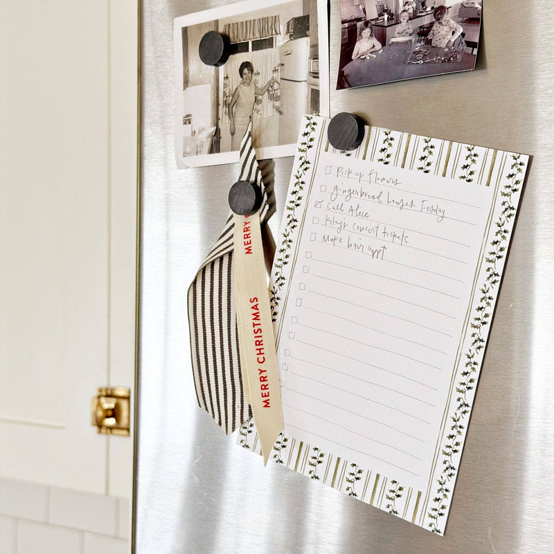 A lined notepad with a decorative border of green leaves and gold stripes, laid flat on a white surface with holiday-themed props around it.