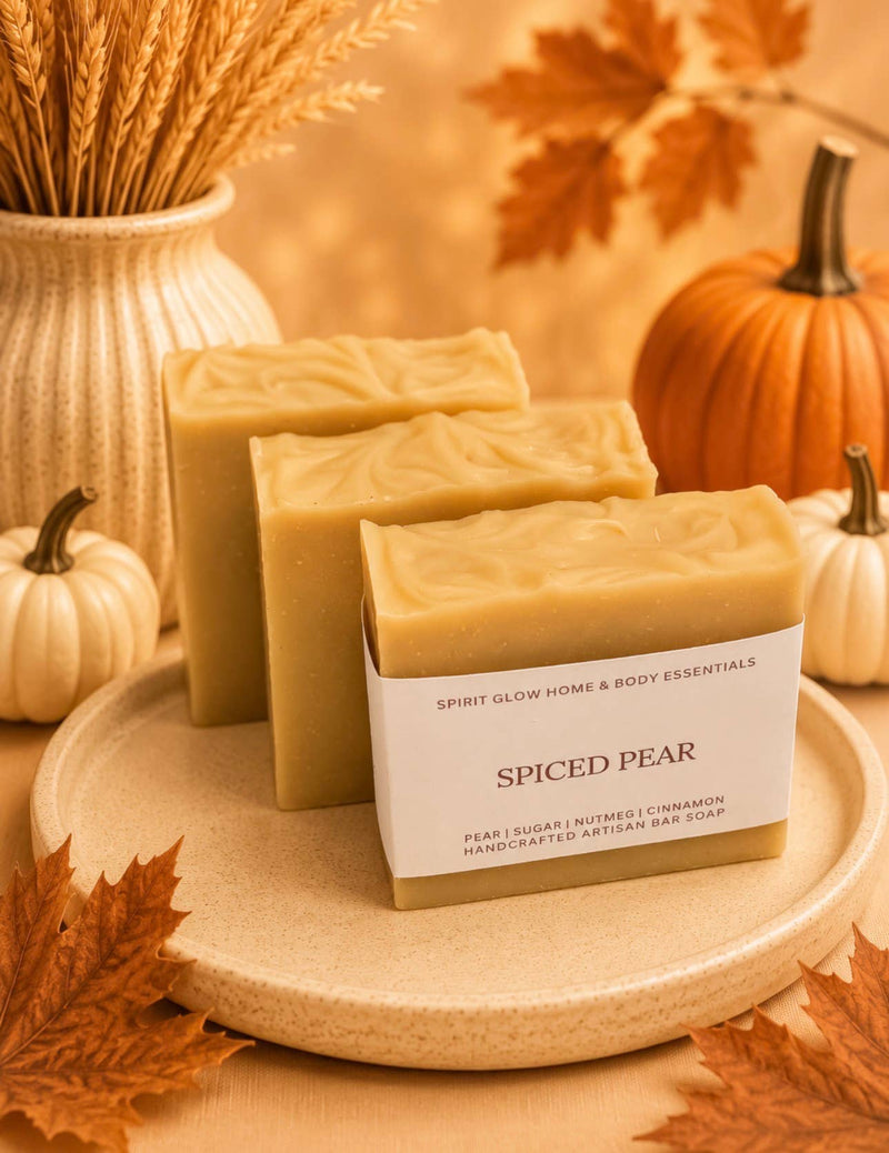 A bar of handmade artisanal bar soap labeled 'SPICED PEAR' by Spirit Glow Home & Body Essentials, displayed on a beige ceramic plate with autumn-themed props including pumpkins, wheat, and leaves.